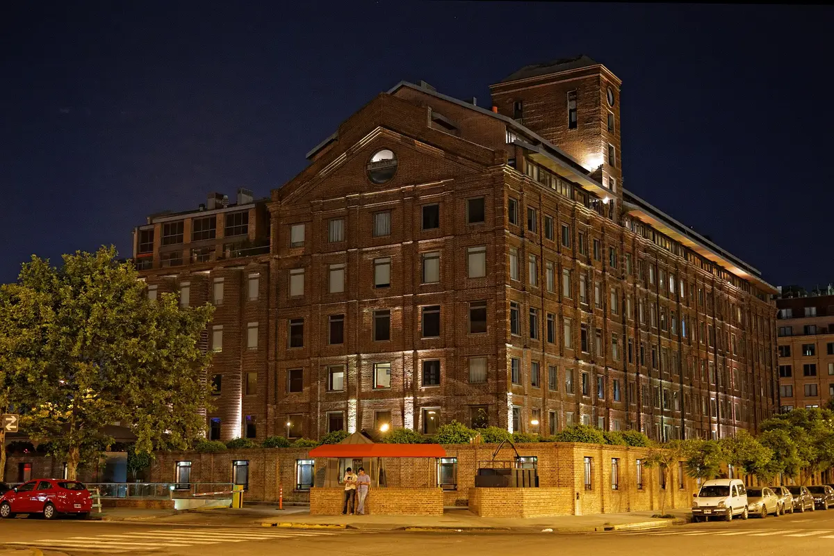 The Faena Hotel in Puerto Madero at night, a lit red-brick former grain mill with a square corner tower, above a low brick wall and a quiet street