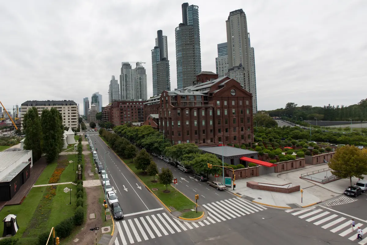 The red-brick Faena Hotel beside a wide tree-lined avenue in Puerto Madero, with glass residential towers rising behind it under a grey sky